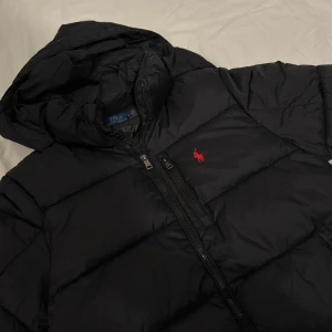 Svart pufferjacka från Polo Ralph Lauren - Svart pufferjacka från Polo Ralph Lauren med huva och tvåvägsdragkedja. Jackan har det klassiska röda Polo-logot broderat på bröstet och flera praktiska fickor med dragkedja. Perfekt för kalla dagar och riktigt snygg stil.