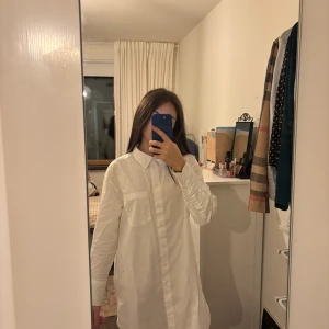 Vit lång skjorta med ficka - Säljer en vit lång skjorta med klassisk krage och bröstficka. Skjortan har en rak och loose passform, långa ärmar och knäppning framtill. Perfekt för en clean och stilren look. Materialet känns som bomull och är lätt och bekvämt.