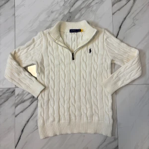 Vit kabelstickad tröja Polo Ralph Lauren - Vit kabelstickad tröja från Polo Ralph Lauren med half zip och broderad logga på bröstet. Tröjan har ribbade muddar och krage, tillverkad i mjuk bomull. Perfekt för dig som gillar klassisk och clean stil.