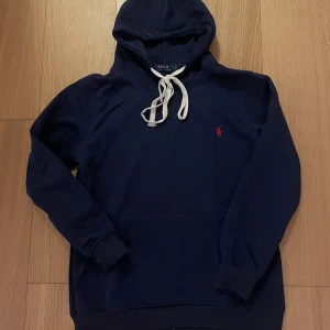 Mörkblå hoodie från Polo Ralph Lauren - Snygg mörkblå hoodie från Polo Ralph Lauren med vit snörning och klassisk röd logobrodyr på bröstet. Tröjan har huva, långa ärmar och ribbade muddar. Perfekt för chill dagar och enkel att matcha med jeans eller joggers.