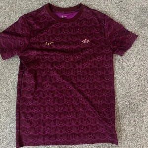 Vinröd Nike KM-mönstrad t-shirt - Säljer en vinröd t-shirt från Nike med unikt grafiskt KM-mönster över hela tröjan. T-shirten har rund halsringning, korta ärmar och små detaljer i gult på bröstet. Perfekt för dig som gillar sportig och stilren design.