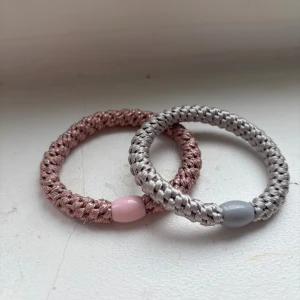 Två flätade armband (Kknekki) - Säljer två snygga, flätade armband i rosa och silver. Armbanden är runda och har en glansig finish med matchande pärldetaljer i plast, en i ljusrosa och en i grått. Perfekta för att ge din outfit en cool och trendig touch.