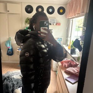 Svart pufferjacka från Colmar - Säljer en svart pufferjacka från Colmar Originals med huva och diskret logga på ärmen. Jackan har en glansig finish, dragkedja framtill och är vadderad för extra värme. Perfekt för dig som vill ha en clean och stilren look under kalla dagar.