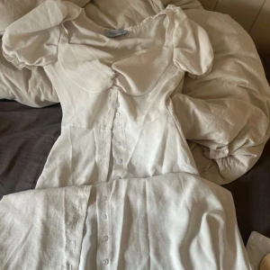 Vit långklänning från Thassity - Superfin vit långklänning från Thassity med puffärmar och markerad byst. Klänningen har knappar hela vägen framtill och är sydd i ett mjukt, lätt material som känns luftigt. Perfekt för dig som gillar romantisk och stilren look.