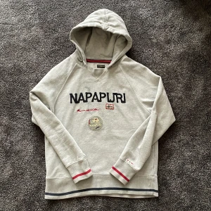 Grå hoodie från Napapijri med brodyr - Grå hoodie från Napapijri med broderad logga, patch och norska flaggan framtill. Muddar med röda detaljer och huva. Skön passform och klassisk streetstil, perfekt för dig som gillar en avslappnad stil. En liten svart prick på ryggen jag inte lyckats få bort tyvärr men går säkert att få bort på något sätt.
