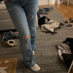 Ljusblå bootcut jeans med slitningar - Säljer ett par ljusblå jeans med bootcut-modell och snygga slitningar på knäna. Jeansen har klassisk femficksdesign, låg midja och är tillverkade i mjukt denimtyg. Perfekta för en avslappnad och trendig look.