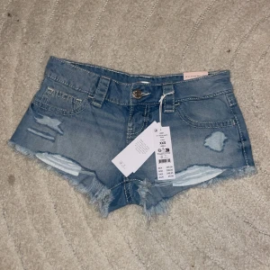 Ljusblå extra lågmidjade jeansshorts - Säljer ett par ljusblå extra lågmidjade jeansshorts från Gina Tricot Young, modell 14+. Shortsen har slitna detaljer, fransad kant och klassiska fem fickor. Perfekt för dig som gillar en chill och trendig look. Passar dig som vill ha riktigt korta shorts med cool vibe. Aldrig andvända.