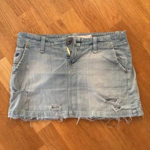 Ljusblå jeanskjol från Nina Jeans - Säljer en cool ljusblå jeanskjol från Nina Jeans med slitna detaljer och rå kant. Kjolen har fem fickor och en knapp- och dragkedjestängning framtill. Perfekt för en avslappnad stil!