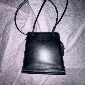 Svart och silver axelväska från Prada - Snygg axelväska från Prada i svart och metallic silver skinn med minimalistisk, fyrkantig form och långa smala axelremmar. Insidan har mönstrat foder med Prada-logga och en praktisk innerficka med dragkedja. Perfekt för dig som gillar stilrena designer. Vintage säljs inte längre den är från 1970 talet någonstans där.