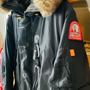 Svart Parajumpers parkajacka med päls - Säljer en svart Parajumpers parkajacka i slim fit med fet päls på huvan, coola detaljer som patch på ärmen, dragkedja och knappar framtill samt flera fickor. Jackan har lång ärm och är perfekt för kalla vinterdagar. Riktigt snygg och funktionell design. Den är en Herrjacka men är snygg på oss kvinnor också därav Unisex. Storlek S men är MKT större i storlek mer M/L. Nypris var 11000kr så detta är ett fynd nu till vintern. Är i mkt gott skick!