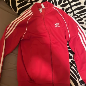 Röd Adidas vindjacka med vita ränder - Säljer en röd Adidas vindjacka med klassiska vita ränder längs ärmarna och Adidas-logga på bröstet. Jackan har dragkedja framtill, ribbad krage och muddar. Perfekt för dig som gillar sportig och retro stil.