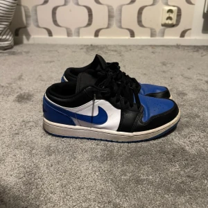 Nike Air Jordan 1 Low ”Royal Toe” - Säljer ett par Nike Air Jordan 1 Low ”Royal Toe”  sneakers med blå, svart och vit färg. Skorna har klassisk snörning, rund tå och platt sula. Ikonisk Jordan-logga på hälen och blå swoosh på sidan. Tillverkade i skinn med syntetdetaljer för extra komfort och stil.