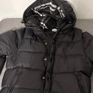 Svart pufferjacka från Burberry - Svart pufferjacka från Burberry med huva och dragkedja. Jackan har ett rutigt foder i huvan och är tillverkad i polyamid och polyester. Klassisk quiltad look med praktiska fickor och snygga detaljer. Perfekt för kalla dagar.