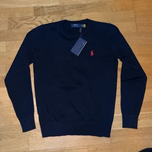 Mörkblå tröja från Polo Ralph Lauren - Snygg mörkblå tröja från Polo Ralph Lauren med klassisk rund halsringning och röd broderad logga på bröstet. Tröjan har långa ärmar och ribbade muddar vid ärmslut och nederkant. Perfekt för en stilren look.