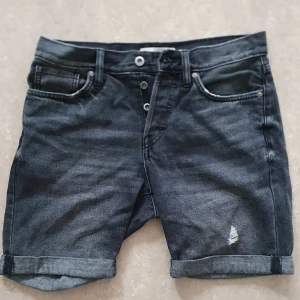 Jeansshorts - Snygga svarta jeansshorts från H&M med upprullade benslut och slitning på ena benet. Klassisk femficksmodell, knappgylf och normal passform. Perfekta för en chill sommarstil.