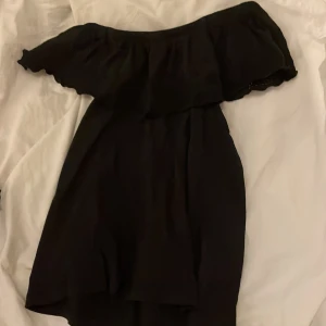 Svart offshoulder blus Lindex - Svart kortklänning från Lindex med offshoulder-modell och bred volang upptill. Volangen har broderade blommor och vågig kant. Klänningen är i mjukt material och har en luftig passform, perfekt för varma dagar.