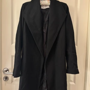 Svart kappa från Selected Femme - Stilren svart kappa från Selected Femme med bred krage och bälte i midjan. Långärmad och med klassisk rak passform, perfekt för dig som gillar minimalistisk look. Materialet är mjukt och har en snygg matt finish. Aldrig använd. Nypris 2199 kr. Hör av er om ni har några funderingar!💗
