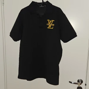 Svart pikétröja - Svart pikétröja från i storlek XXXL med gul broderad logga på bröstet. Klassisk krage och korta ärmar, tillverkad i ett strukturerat material som känns lyxigt. Perfekt för dig som gillar exklusiva märken och vill sticka ut med en fet logga. Skulle säga att den sitter mer som S/M