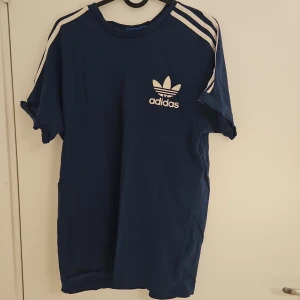Blå Adidas t-shirt med vita ränder - Snygg blå t-shirt från Adidas med klassiska vita ränder på axlarna och det ikoniska Adidas-trycket på bröstet. T-shirten har rund hals och korta ärmar, tillverkad i mjuk bomull som är skön mot huden. Perfekt för dig som gillar sportig och avslappnad stil.