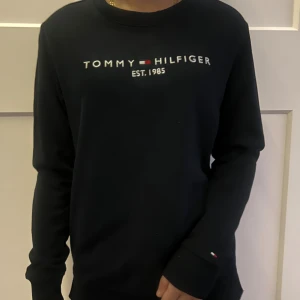 Svart sweatshirt från Tommy Hilfiger - Svart sweatshirt från Tommy Hilfiger med broderad logga och text 'EST. 1985' på bröstet. Klassisk rund hals, långa ärmar och ribbade muddar. Detalj med Hilfiger-flagga på ärmslut och insida slits. Mjuk bomullskänsla och relaxed fit. Köpt i USA men kommer inte till användning. Det går att buda på❤️