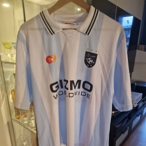 Randig fotbollströja Gizmo Worldwide - Snygg fotbollströja från Gizmo med ljusblå och vita ränder, svart tryck och klubbmärke på bröstet. Klassisk krage med svarta ränder och nummer 23 på ryggen. Tillverkad i lätt och ventilerande meshmaterial, perfekt för match eller träning.