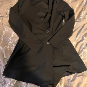 Svart kort klänning med kavajlook - Stilren svart kort klänning med kavajskärning och långa ärmar. Klänningen har djup v-ringning, två knappar framtill och markerad midja. Ser ut som en kjol framifrån men som shorts bakifrån. Storlek 40 men passar även 36/38