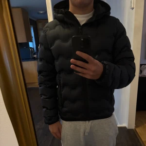 Svart pufferjacka från Colmar - Säljer en svart pufferjacka från Colmar med huva och snyggt vågformat quiltat mönster. Jackan har dragkedja framtill och två sidofickor med dragkedja. Perfekt för dig som vill ha en stilren och varm jacka med modern look.