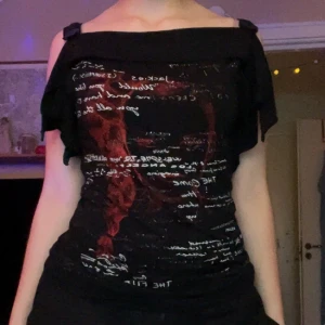 Svart offshoulder topp med tryck - Cool svart offshoulder topp från YRC med dekorativa knappar på axlarna. Framsidan har ett stort grafiskt tryck med röd illustration av ett ansikte och massa vit text. Materialet är mjukt och stretchigt, perfekt för dig som gillar edgy och unik stil. Storlek S/M.