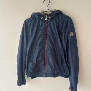 Marinblå vindjacka från Moncler - Snygg marinblå vindjacka från Moncler med huva och grått innerfoder. Jackan har dragkedja med röd och vit kant, två fickor och Moncler-logga på ärmen. Perfekt för dig som gillar stilrena och sportiga plagg med premiumkänsla.