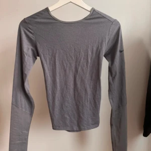 Grå långärmad träningströja från NIKE - Grå långärmad träningströja från Nike i storlek S. Lätt, stretchigt och snabbtorkande material. Baksidan har en snygg omlott- och öppningsdetalj. Figurnära passform. Använd fåtal gånger och är därför i bra och fin kvalité.