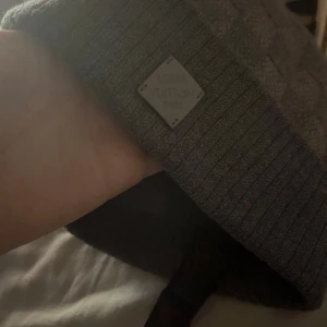 Grå Louis Vuitton mössa - Snygg grå mössa från Louis Vuitton med diskret logoplatta framtill. Stickad i mjukt material med ribbad kant och klassisk form. Perfekt för dig som vill ha en lyxig och stilren accessoar till höst och vinter.