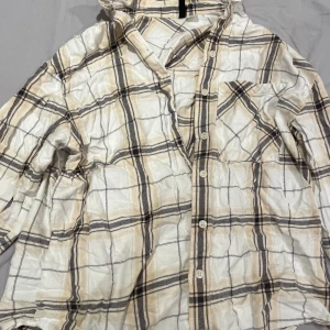 Beige och vit rutig skjorta - Rutig skjorta i beige, vitt och svarta toner. Skjortan har knappar framtill, lång ärm och en bröstficka. Har en avslappnad passform. Perfekt för en chill och stilren look.