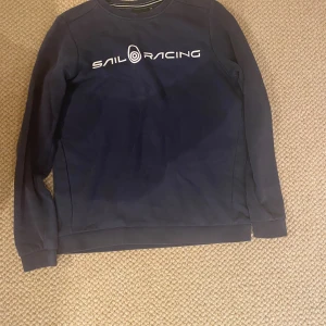 Marinblå Sail Racing tröja - Säljer en marinblå långärmad tröja från Sail Racing med vit logga tryckt över bröstet. Tröjan har rund halsringning och är tillverkad i ett mjukt material som passar perfekt för kyligare dagar. Det är nästan hål på en av ärmarnas armbåge. Hör av dig vid frågor o funderingar