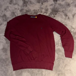 Vinröd tröja från Polo Ralph Lauren - Vinröd stickad tröja från Polo Ralph Lauren i storlek XXL. Klassisk rund halsringning och diskret broderad logga på bröstet. Tröjan har ribbade muddar vid ärmslut och nederkant, perfekt för lager-på-lager under kyliga dagar.