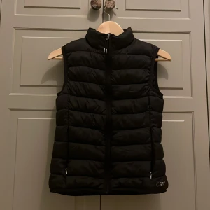 Svart dunväst från CRW - Svart dunväst från CRW med hög krage och dragkedja framtill. Västen har två sidofickor med dragkedja och quiltad design. Perfekt för lager-på-lager under kyliga dagar.