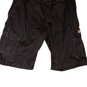 Svarta cargoshorts från Björn kläder  - Svarta cargoshorts från Hydroscand med kontrastsömmar och flera praktiska fickor. Märke Björkläder, tillverkade i en slitstark mix av polyester och bomull. Perfekta för dig som gillar funktionella och stilrena shorts med streetkänsla.