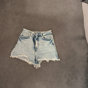 Ljusblåa high waist jeansshorts - Säljer ett par ljusblåa denimshorts med hög midja och råa, fransiga kanter. Klassisk femficksmodell med bälteshällor och dragkedja fram. 