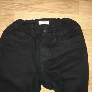 Svarta jeansbyxor från Lindex - Svarta jeansbyxor från Lindex med klassisk femficksdesign och knappstängning. Byxorna har rak passform och elastisk midja för extra komfort. Perfekt för dig som gillar stilrena och enkla jeans i svart.