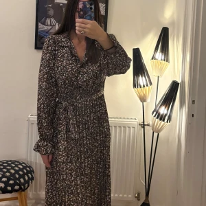 Blommig långklänning från Hapsho - Supersöt långklänning från Hapsho med småblommigt mönster i beige, rosa och vitt på svart botten. Klänningen har långa ärmar, knytband i midjan och klassisk krage. Perfekt för dig som gillar romantisk och avslappnad stil.