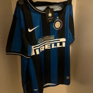 Inter Milan randig fotbollströja Nike - Säljer en officiell Inter Milan fotbollströja från Nike med Dri-FIT teknologi. Tröjan är blå och svart med breda ränder, korta ärmar och broderat klubbmärke på bröstet. Pirelli-logga på framsidan och detaljer om Champions League-finalen 2010. Materialet är lätt och andas.