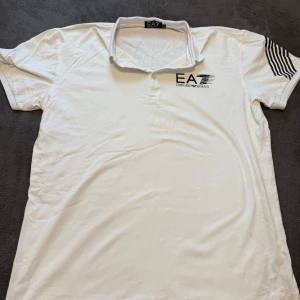 Vit EA7 Emporio Armani piké XXL - Snygg vit pikétröja från EA7 Emporio Armani i storlek XXL. Tröjan har svart logotyptryck på bröstet och coola svarta ränder på ena ärmen. Klassisk krage och korta ärmar, tillverkad i mjuk bomull som är skön mot huden. Perfekt för dig som gillar stilrena märkeskläder.