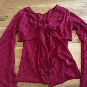 Vinröd omlottopp med knyt från SHEIN - Säljer en vinröd omlottopp från SHEIN i storlek M. Toppen har långa ärmar, snygg knytning framtill och ett vågigt, ribbat mönster i tyget. Materialet är lätt och syntetiskt, perfekt för att skapa en cool och trendig look.