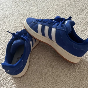 Blå adidas Campus sneakers - Snygga blå adidas Campus sneakers med klassiska vita ränder på sidorna och vit sula med gummiyttersula. Ovandelen är i mocka och skorna har snörning. Perfekta för dig som gillar retrostil och streetwear.