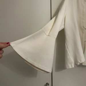 Vit blazer/kofta byTiMo - Säljer en stilren vit trekvartsärmad kofta från byTiMo med vida trumpetärmar och öppen front. Toppen har en rak passform och är tillverkad i ett mjukt tyg som känns skönt mot huden. Perfekt att slänga över en klänning för en clean look. Tror inte den säljs längre