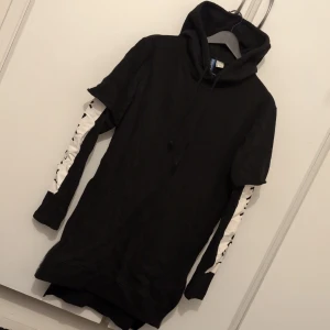 Svart hoodie med japansk text - Svart lång hoodie med huva och snörning, vita paneler med japansk text längs ärmarna och dragkedjor i sidorna. Oversized passform och stor ficka framtill. S men stor i storleken!