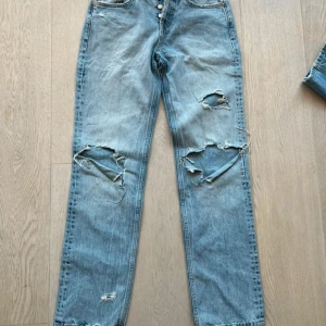 Ljusblå ripped jeans från Zara - Säljer ett par ljusblå jeans från Zara i storlek 36. Jeansen har raka ben, mid midja och slitna detaljer med stora hål på båda benen. Klassisk femficksmodell och tillverkade i bomull. Perfekta för en avslappnad och trendig look.