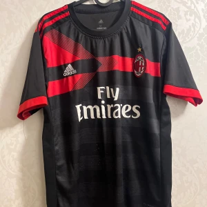 AC Milan Adidas fotbollströja XL - Snygg AC Milan fotbollströja från Adidas i svart med röda detaljer och ränder på axlarna. Tryck med Fly Emirates och klubbmärke på bröstet. Kortärmad modell i lätt och ventilerande polyester, perfekt för match eller träning.