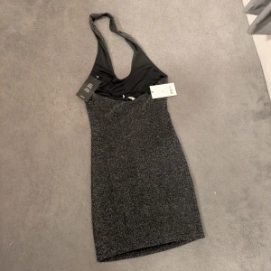 Glittrig svart kortklänning  - Supersnygg svart glittrig kortklänning med halterneck och djup urringning. Tight passform som framhäver figuren och perfekt för att sticka ut. Materialet är stretchigt och skimrar snyggt i ljuset. 