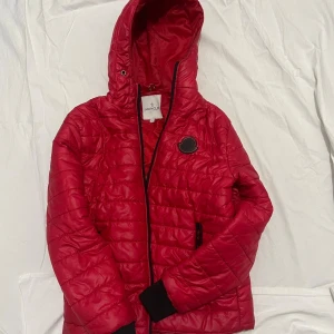 Röd pufferjacka från Moncler - Säljer en röd pufferjacka från Moncler med huva och svart Moncler-logga på bröstet. Jackan har quiltad design, svarta muddar vid ärmslut och dragkedja framtill. Insidan har en cool serietidningsetikett och flera detaljer. Perfekt för dig som vill sticka ut.
