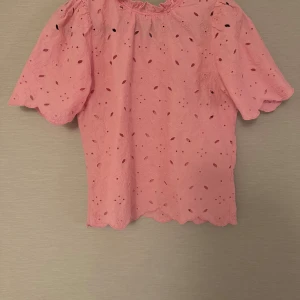 Rosa blus med broderade hål och volang - Supersöt rosa blus med broderade blommor och hål, puffiga korta ärmar och volangkant nertill. Blusen har rund halsringning och är lätt och luftig.